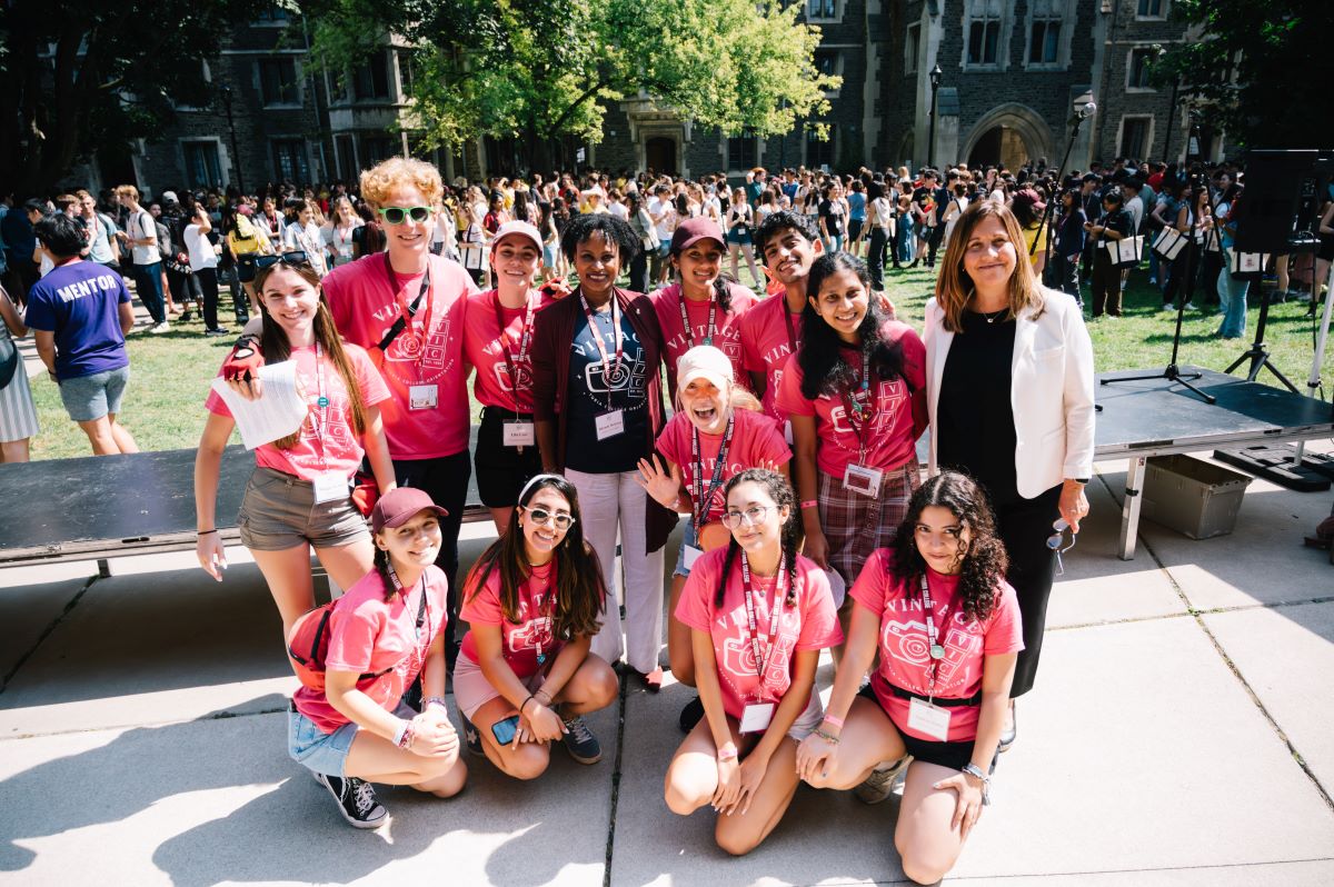 A group of smiling students wearing red orientation t-shirts pose for photos with university staff.