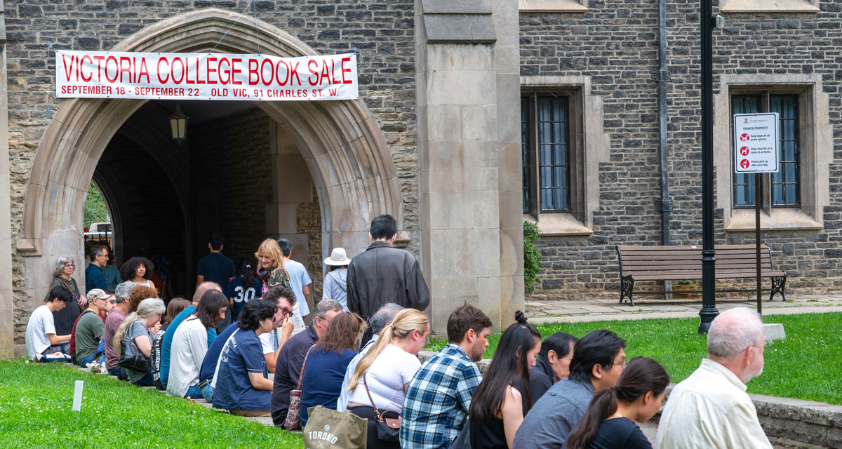 Many book collectors seated along Burwash Quad near an archway, with a Victoria College Book Sale sign hanging above in the background.