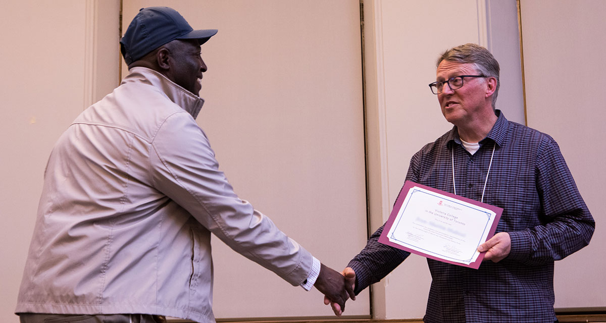 Two men share a congratulatory handshake as one is awarded a diploma during a Humanities for Humanity event at Alumni Hall, Victoria University, University of Toronto.